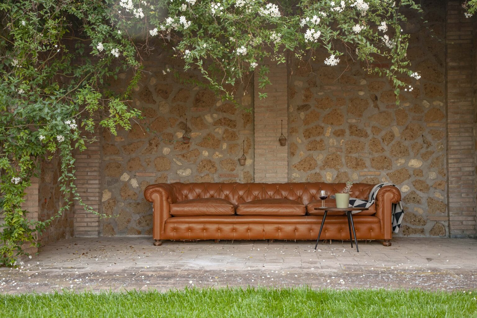 Cozy outdoor seating area featuring a stylish brown leather Chesterfield sofa, a small coffee table with a plant and wine glass, surrounded by lush greenery and a rustic stone wall. | Only on SopranoVillas