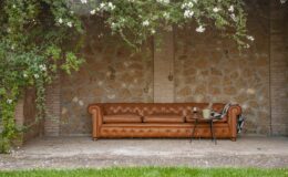 Cozy outdoor seating area featuring a stylish brown leather Chesterfield sofa, a small coffee table with a plant and wine glass, surrounded by lush greenery and a rustic stone wall.