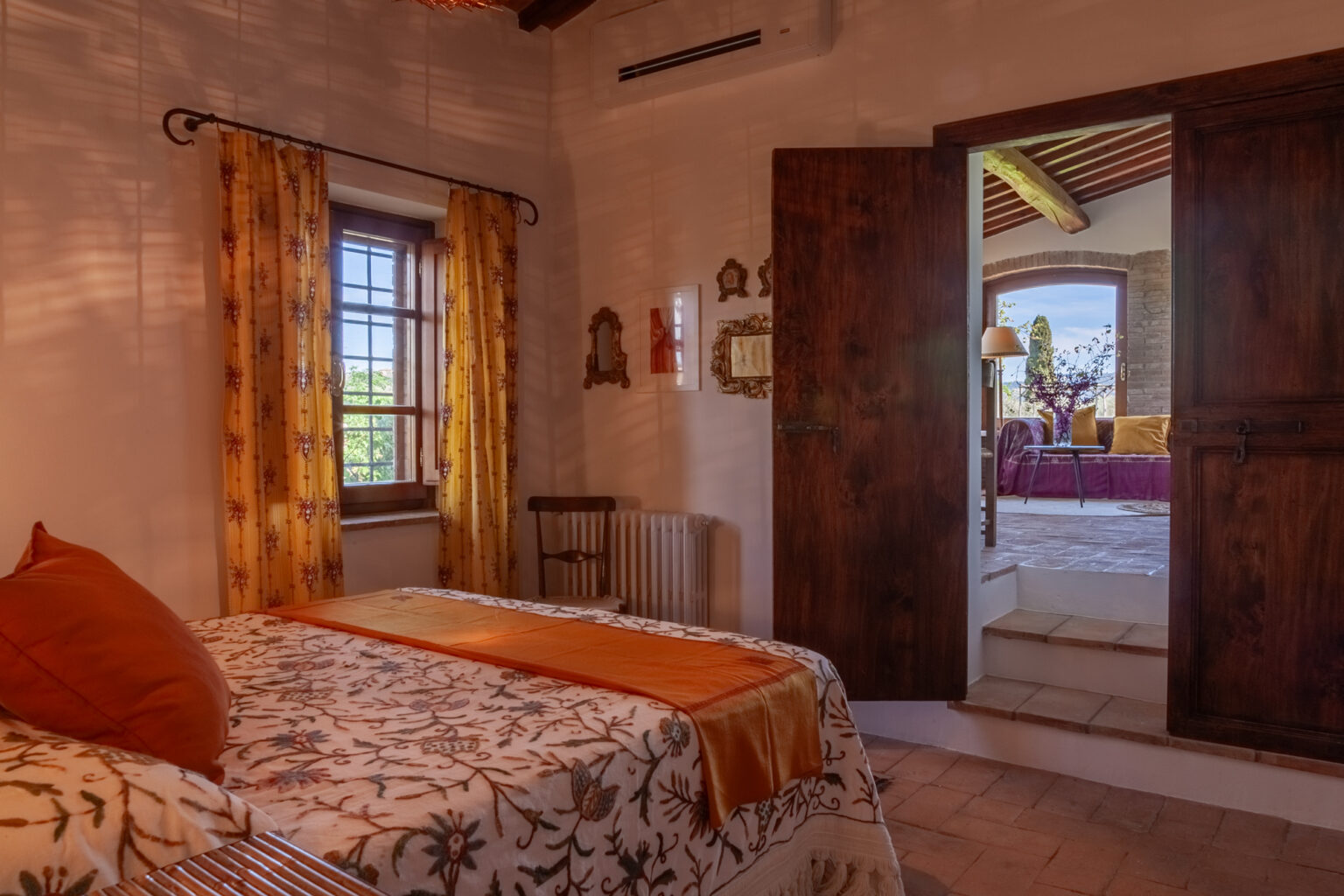 Cozy bedroom featuring a floral-patterned bedspread, orange accents, and yellow curtains. A wooden chair and decorative mirrors adorn the walls. The doorway leads to a bright living area with a view of the outdoors, enhancing the inviting atmosphere. | Only on SopranoVillas