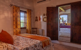 Cozy bedroom featuring a floral-patterned bedspread, orange accents, and yellow curtains. A wooden chair and decorative mirrors adorn the walls. The doorway leads to a bright living area with a view of the outdoors, enhancing the inviting atmosphere.