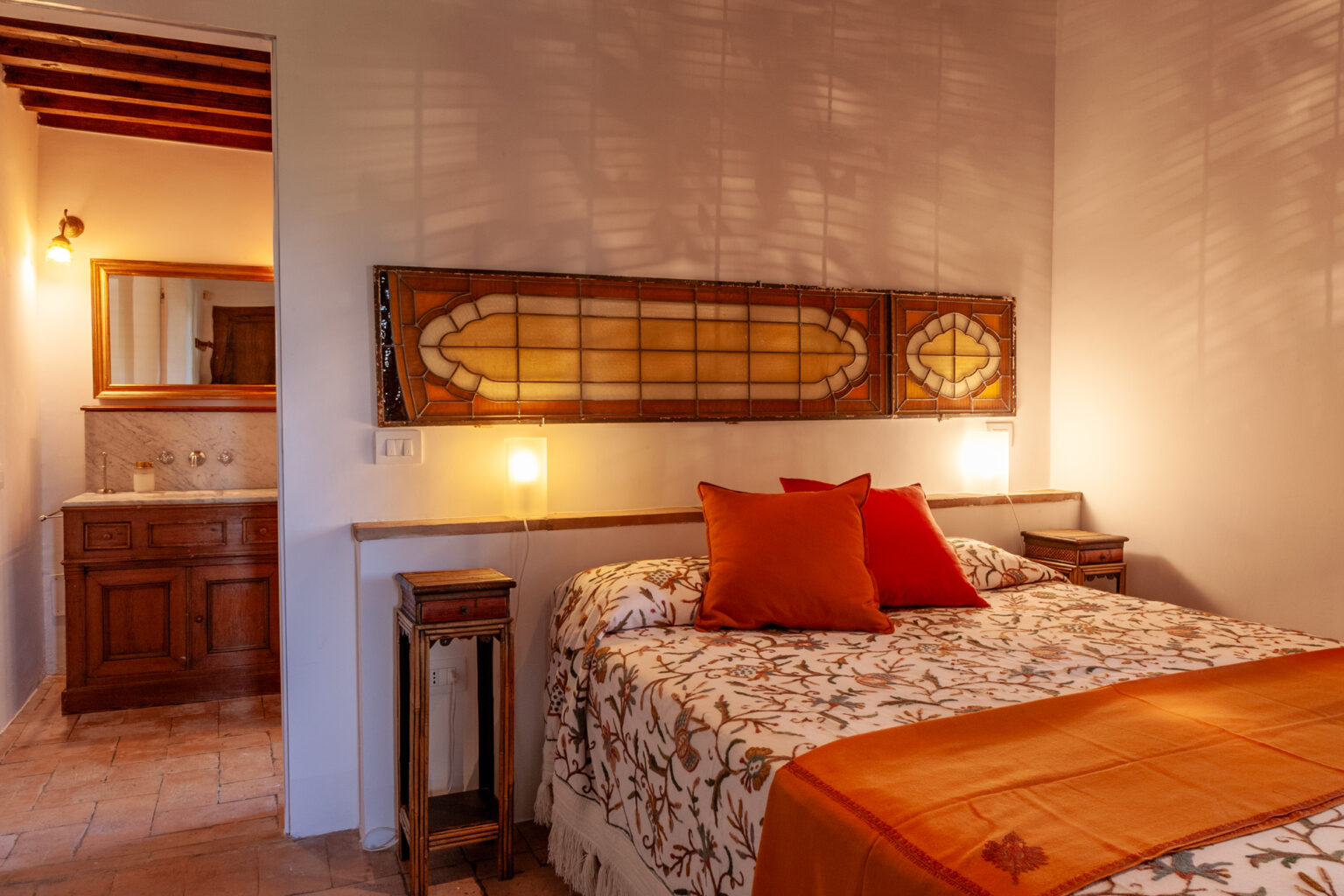 Cozy bedroom interior featuring a queen-sized bed with decorative pillows, warm lighting, and wooden accents, complemented by a stylish bathroom area in the background. Ideal for a relaxing stay. | Only on SopranoVillas