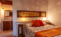 Cozy bedroom interior featuring a queen-sized bed with decorative pillows, warm lighting, and wooden accents, complemented by a stylish bathroom area in the background. Ideal for a relaxing stay.