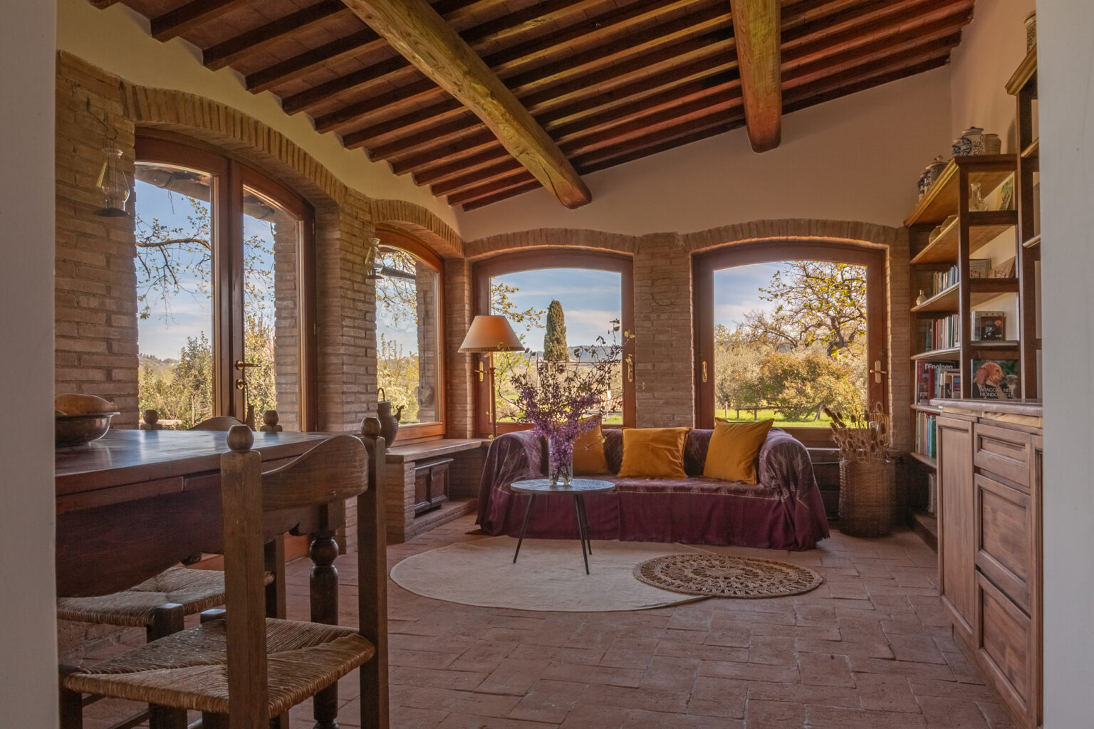 Cozy living room with large windows showcasing a scenic view, featuring a maroon sofa with decorative cushions, a wooden dining table, and a bookshelf against a rustic brick wall, all under a wooden beam ceiling. | Only on SopranoVillas