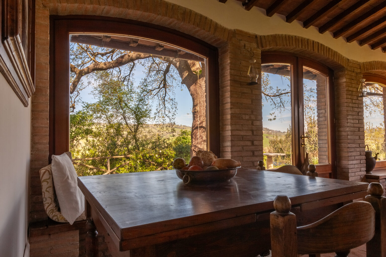 Cozy dining area with a wooden table, featuring a bowl of fruit, and large windows offering a scenic view of lush greenery and trees outside. | Only on SopranoVillas