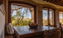 Cozy dining area with a wooden table, featuring a bowl of fruit, and large windows offering a scenic view of lush greenery and trees outside.