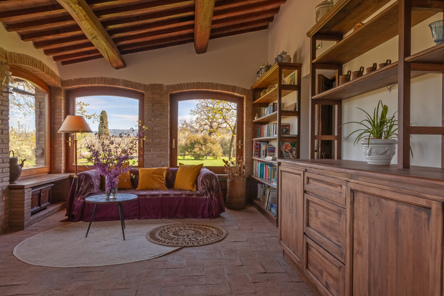 Cozy living room with rustic wooden beams, large windows showcasing a scenic view, a comfortable sofa adorned with colorful cushions, and a wooden bookshelf filled with decorative items and plants. | Only on SopranoVillas