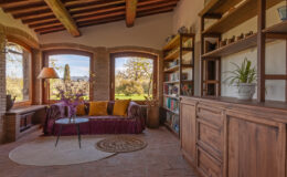 Cozy living room with rustic wooden beams, large windows showcasing a scenic view, a comfortable sofa adorned with colorful cushions, and a wooden bookshelf filled with decorative items and plants.