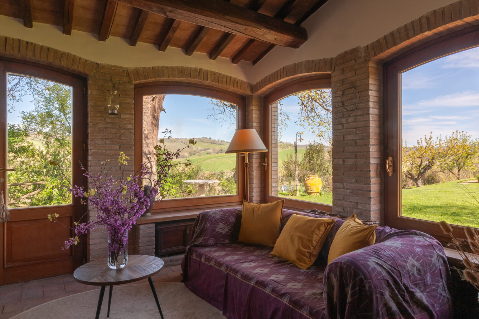 Cozy sunroom with large windows offering a scenic view of greenery, featuring a plush sofa adorned with colorful cushions and a decorative vase of flowers. Ideal for relaxation and enjoying natural light. | Only on SopranoVillas