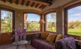 Cozy sunroom with large windows offering a scenic view of greenery, featuring a plush sofa adorned with colorful cushions and a decorative vase of flowers. Ideal for relaxation and enjoying natural light.