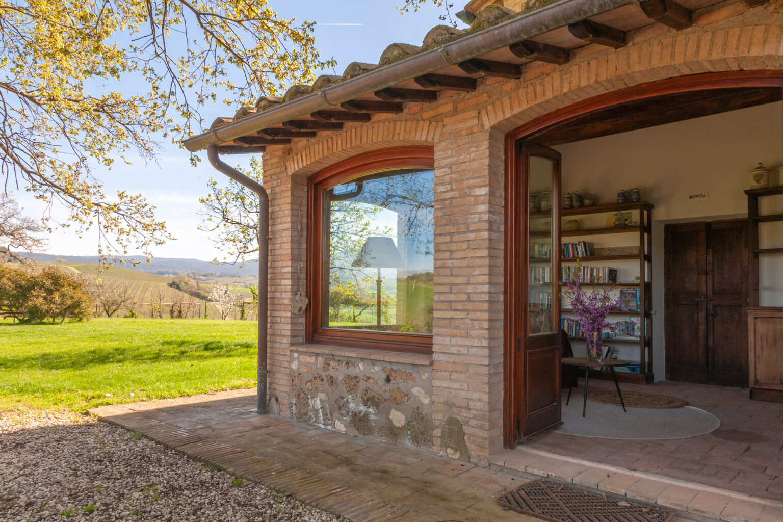 Cozy rustic building with large windows, featuring a bookshelf and a chair, surrounded by lush green landscape and trees, ideal for a tranquil retreat or workspace. | Only on SopranoVillas