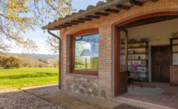 Cozy rustic building with large windows, featuring a bookshelf and a chair, surrounded by lush green landscape and trees, ideal for a tranquil retreat or workspace.