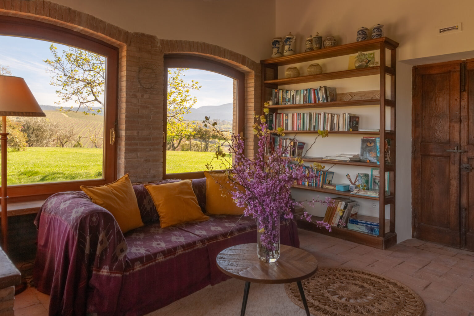Cozy living room with large windows offering a view of greenery, featuring a comfortable sofa adorned with yellow cushions, a coffee table, a bookshelf filled with books, and a vase of fresh flowers. | Only on SopranoVillas