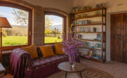 Cozy living room with large windows offering a view of greenery, featuring a comfortable sofa adorned with yellow cushions, a coffee table, a bookshelf filled with books, and a vase of fresh flowers.