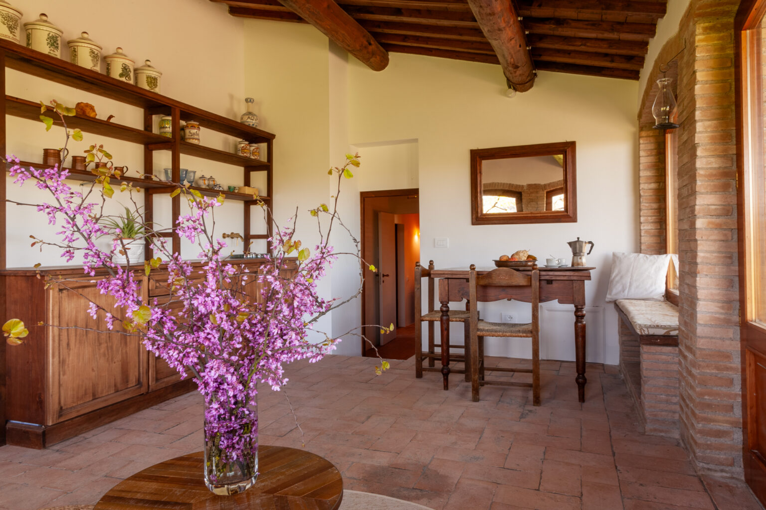 Cozy interior of a rustic room featuring a wooden table and chairs, shelves filled with decorative items, and a vase of pink flowers on a round table, showcasing a warm and inviting atmosphere. | Only on SopranoVillas