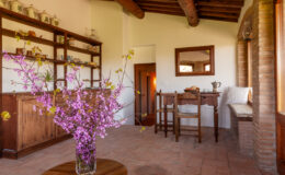 Cozy interior of a rustic room featuring a wooden table and chairs, shelves filled with decorative items, and a vase of pink flowers on a round table, showcasing a warm and inviting atmosphere.