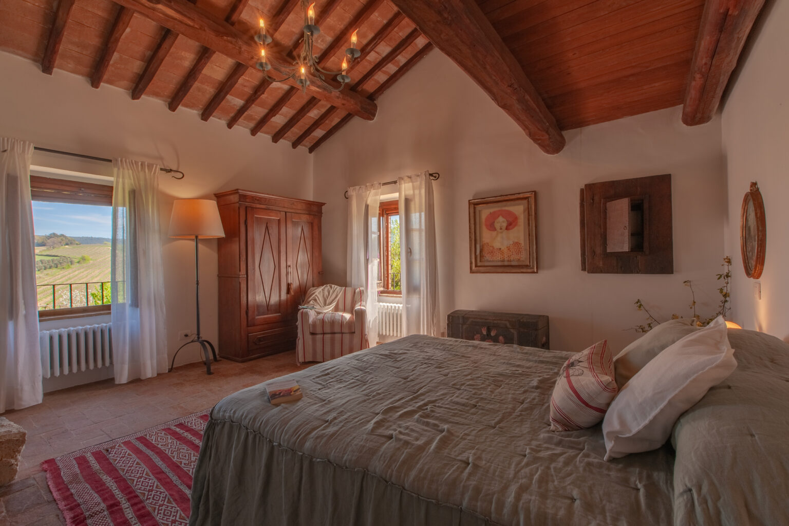 Cozy bedroom featuring a wooden beam ceiling, a comfortable bed with decorative pillows, and a vintage armoire. Natural light streams in through a window with sheer curtains, offering a scenic view of vineyards outside. The room is accented with a stylish lamp, a striped armchair, and framed artwork, creating a warm and inviting atmosphere. | Only on SopranoVillas