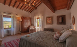 Cozy bedroom featuring a wooden beam ceiling, a comfortable bed with decorative pillows, and a vintage armoire. Natural light streams in through a window with sheer curtains, offering a scenic view of vineyards outside. The room is accented with a stylish lamp, a striped armchair, and framed artwork, creating a warm and inviting atmosphere.