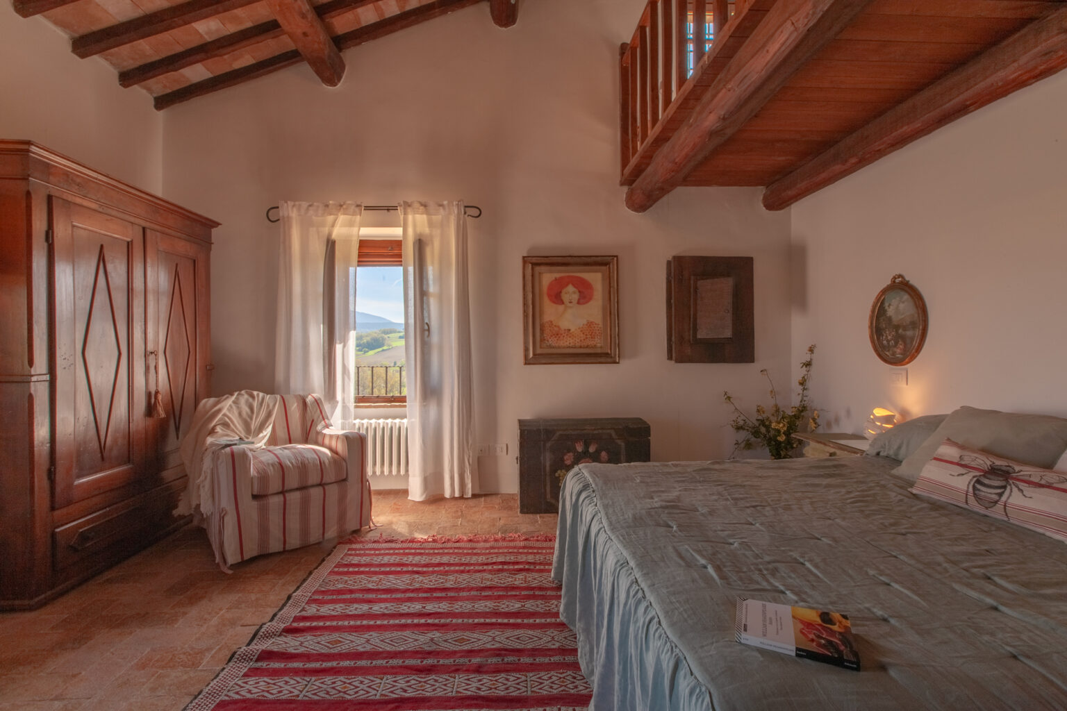 Cozy bedroom with wooden beams, featuring a comfortable bed, vintage wardrobe, and a sitting area by the window, adorned with natural light and scenic views. Decor includes artwork and a patterned rug, creating a warm and inviting atmosphere. | Only on SopranoVillas