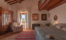Cozy bedroom with wooden beams, featuring a comfortable bed, vintage wardrobe, and a sitting area by the window, adorned with natural light and scenic views. Decor includes artwork and a patterned rug, creating a warm and inviting atmosphere.
