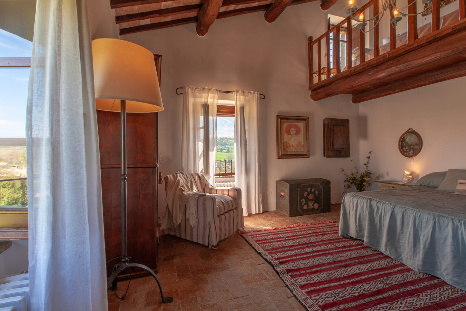 Cozy bedroom with rustic decor, featuring a comfortable bed, a striped area rug, a reading chair, and large windows that provide natural light and scenic views. The room has wooden beams and soft curtains, creating a warm and inviting atmosphere. | Only on SopranoVillas