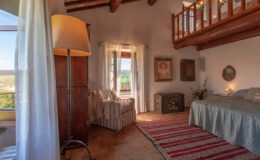 Cozy bedroom with rustic decor, featuring a comfortable bed, a striped area rug, a reading chair, and large windows that provide natural light and scenic views. The room has wooden beams and soft curtains, creating a warm and inviting atmosphere.