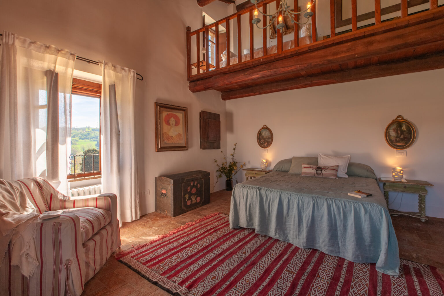 Cozy bedroom with a wooden loft, featuring a large bed with a blue coverlet, a striped area rug, and a comfortable armchair. Natural light streams in through large windows, highlighting decorative wall art and a vintage chest. Ideal for a relaxing retreat. | Only on SopranoVillas