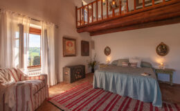 Cozy bedroom with a wooden loft, featuring a large bed with a blue coverlet, a striped area rug, and a comfortable armchair. Natural light streams in through large windows, highlighting decorative wall art and a vintage chest. Ideal for a relaxing retreat.
