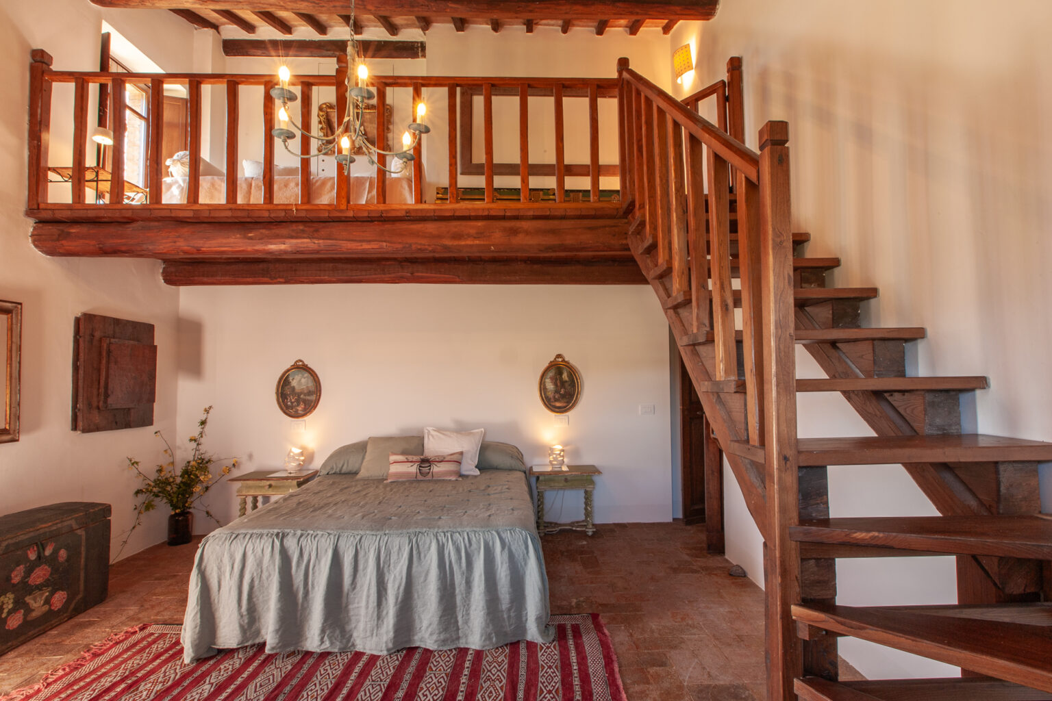 Cozy bedroom interior featuring a wooden loft, a queen-sized bed with a light green bedspread, rustic decor, and a staircase leading to the upper level. Natural light illuminates the space, enhancing the warm atmosphere. | Only on SopranoVillas