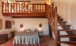 Cozy bedroom interior featuring a wooden loft, a queen-sized bed with a light green bedspread, rustic decor, and a staircase leading to the upper level. Natural light illuminates the space, enhancing the warm atmosphere.