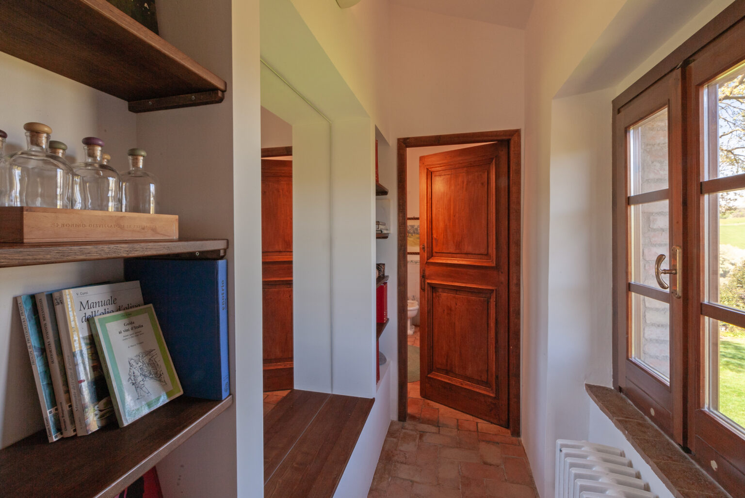 Interior view of a cozy hallway featuring wooden shelves with books and glass bottles, a mirror, and a wooden door leading to another room, complemented by natural light from a nearby window. | Only on SopranoVillas