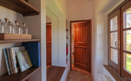 Interior view of a cozy hallway featuring wooden shelves with books and glass bottles, a mirror, and a wooden door leading to another room, complemented by natural light from a nearby window.