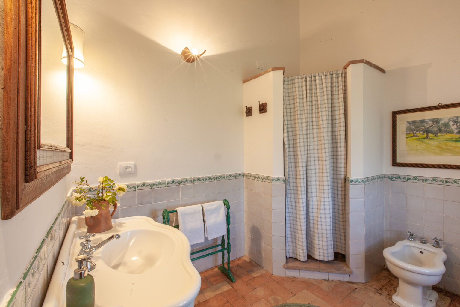 Bright and airy bathroom featuring a shower with a curtain, a sink with a vintage faucet, a bidet, and decorative wall tiles, complemented by natural light and a framed landscape painting. | Only on SopranoVillas