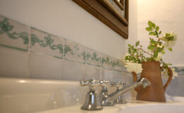 Close-up view of a bathroom sink featuring vintage-style faucets, decorative tiled backsplash, and a clay pitcher with fresh flowers, creating a charming and inviting atmosphere.