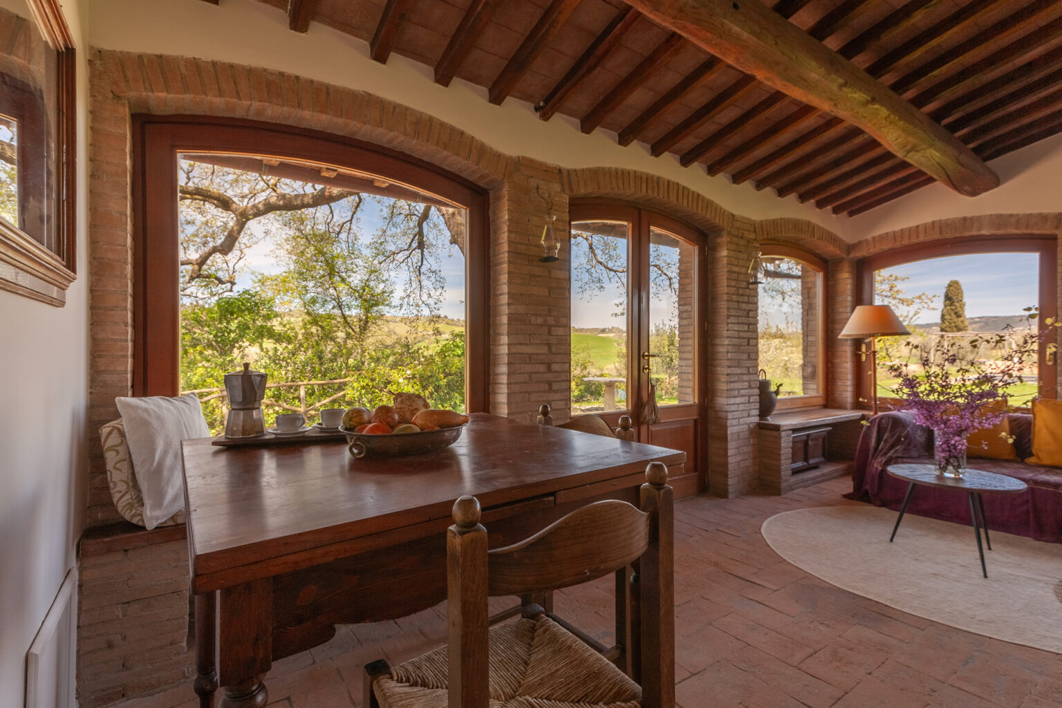 Cozy sunlit room with large windows showcasing scenic views, featuring a wooden dining table with fruit, comfortable seating, and rustic decor. Ideal for relaxation and enjoying nature. | Only on SopranoVillas
