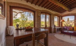 Cozy sunlit room with large windows showcasing scenic views, featuring a wooden dining table with fruit, comfortable seating, and rustic decor. Ideal for relaxation and enjoying nature.