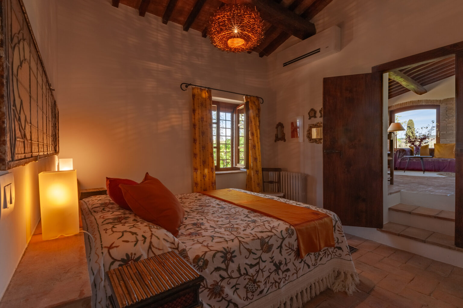 Cozy bedroom featuring a comfortable bed with decorative pillows, warm lighting, and rustic wooden accents. The room has a large window with yellow curtains, providing a view of the outdoors, and opens to a well-lit hallway. | Only on SopranoVillas