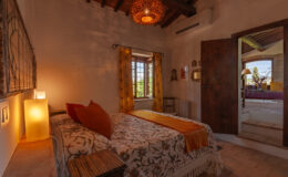 Cozy bedroom featuring a comfortable bed with decorative pillows, warm lighting, and rustic wooden accents. The room has a large window with yellow curtains, providing a view of the outdoors, and opens to a well-lit hallway.