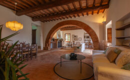 Spacious living room featuring rustic wooden beams, an arched entrance, and a cozy seating area with a coffee table. Natural light illuminates the space, showcasing a blend of modern and traditional decor.