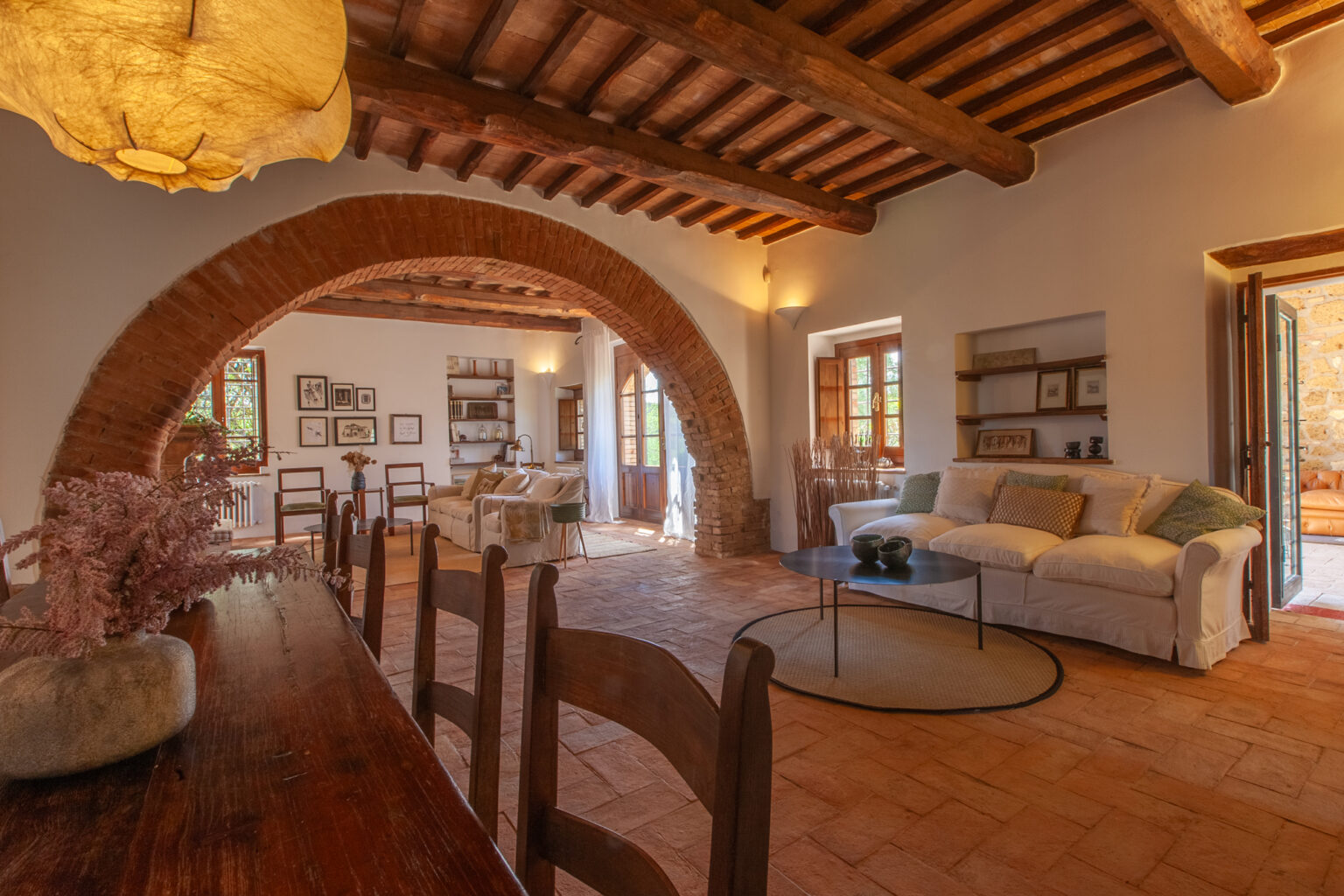 Spacious living area with rustic wooden beams, arched entryways, and a cozy seating arrangement featuring a white sofa. Natural light floods the room through large windows, enhancing the warm ambiance and inviting decor. | Only on SopranoVillas