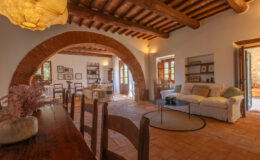 Spacious living area with rustic wooden beams, arched entryways, and a cozy seating arrangement featuring a white sofa. Natural light floods the room through large windows, enhancing the warm ambiance and inviting decor.