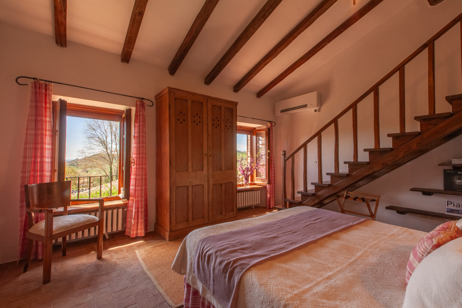 Cozy bedroom interior featuring wooden beams, a double bed with soft linens, a rustic wardrobe, and a chair by the window. Natural light floods the space, highlighting the charming decor and the view of the outdoors through the open window with pink checkered curtains. A staircase leads to the upper level, adding to the room's inviting atmosphere. | Only on SopranoVillas