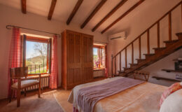 Cozy bedroom interior featuring wooden beams, a double bed with soft linens, a rustic wardrobe, and a chair by the window. Natural light floods the space, highlighting the charming decor and the view of the outdoors through the open window with pink checkered curtains. A staircase leads to the upper level, adding to the room's inviting atmosphere.