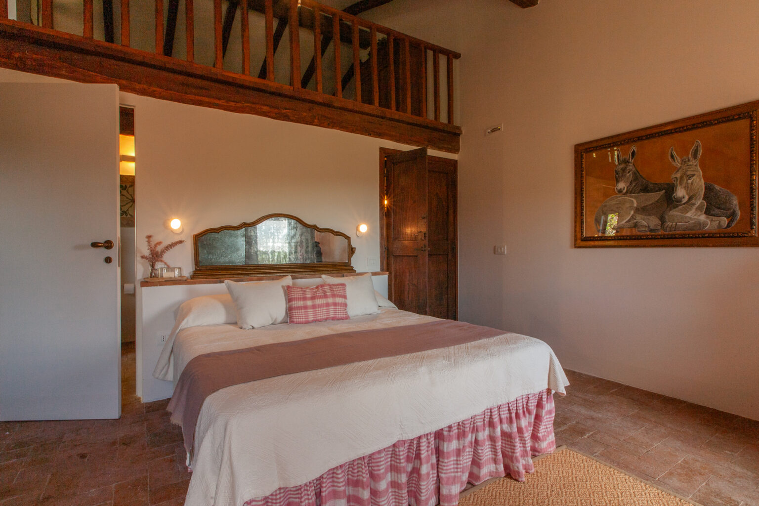 Cozy bedroom featuring a double bed with a pink and white patterned bedspread, decorative pillows, and a vintage mirror. The room has wooden beams, a rustic wooden door, and a framed artwork of donkeys on the wall. Soft lighting creates a warm ambiance, enhancing the inviting atmosphere. | Only on SopranoVillas