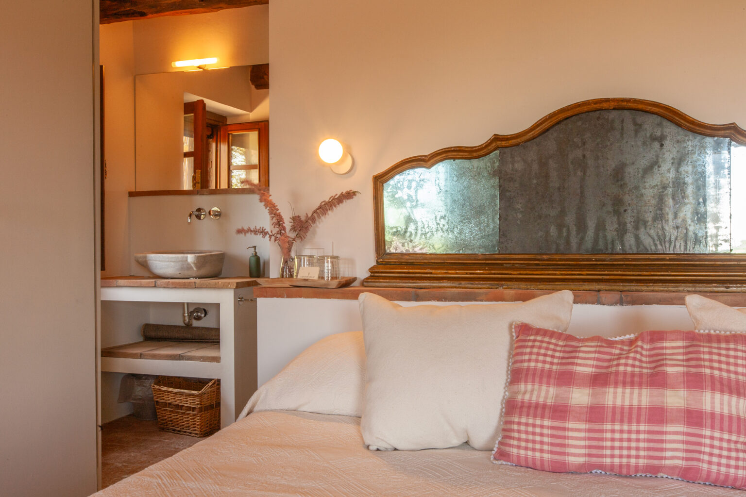 Cozy bedroom interior featuring a soft bed with decorative pillows, a vintage-style mirror, and a stylish bathroom area with a stone sink and natural decor elements. Warm lighting enhances the inviting atmosphere. | Only on SopranoVillas