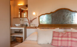 Cozy bedroom interior featuring a soft bed with decorative pillows, a vintage-style mirror, and a stylish bathroom area with a stone sink and natural decor elements. Warm lighting enhances the inviting atmosphere.