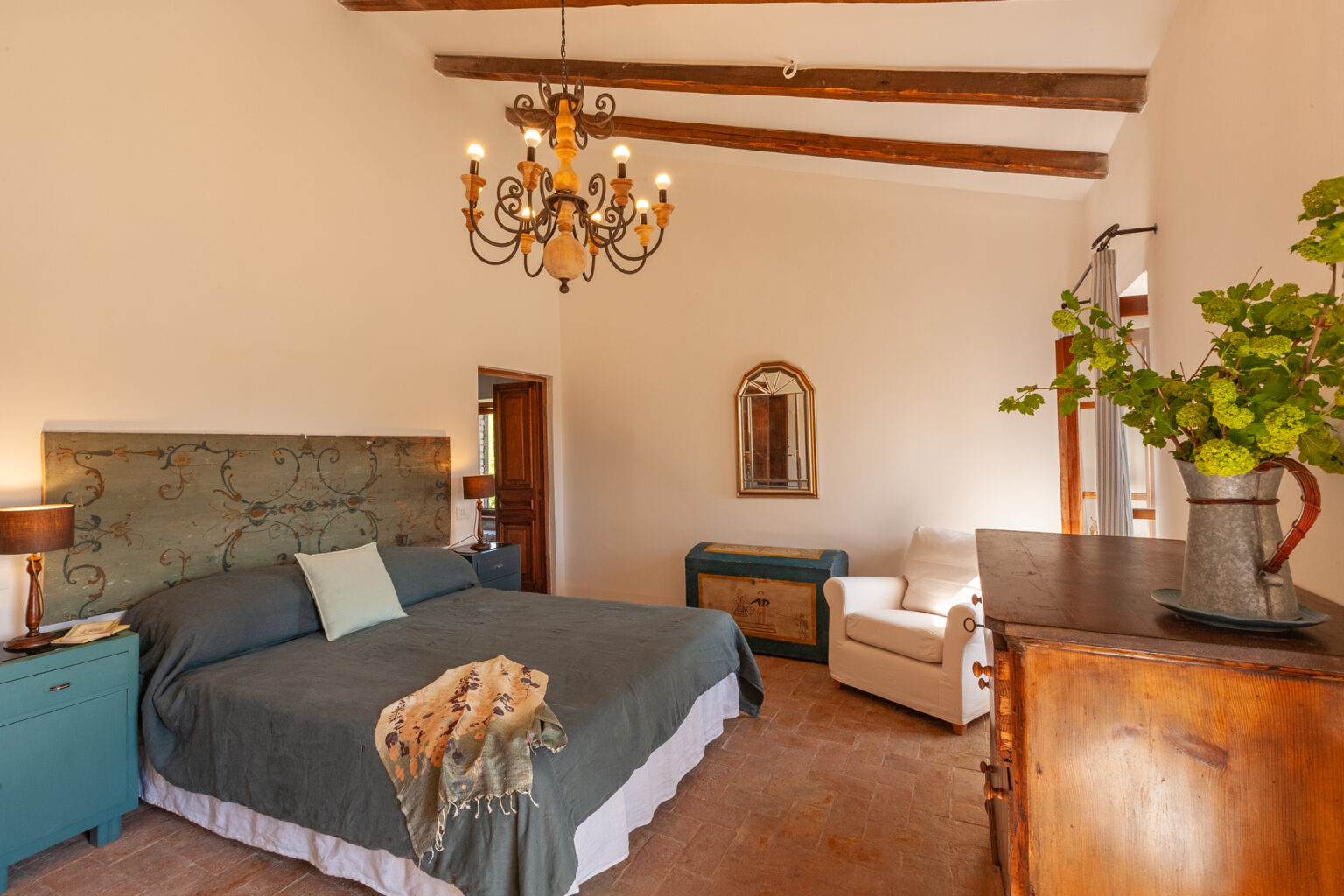 Spacious and elegantly designed bedroom featuring a large bed with decorative throw, a vintage wooden dresser, a comfortable armchair, and a stylish chandelier, complemented by warm lighting and rustic wooden beams. | Only on SopranoVillas