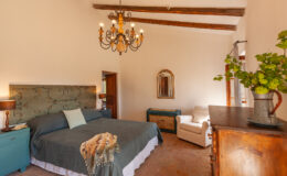 Spacious and elegantly designed bedroom featuring a large bed with decorative throw, a vintage wooden dresser, a comfortable armchair, and a stylish chandelier, complemented by warm lighting and rustic wooden beams.
