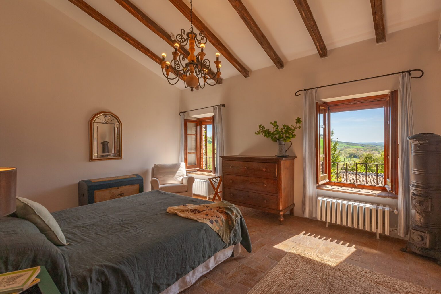 Cozy bedroom featuring a green bedspread, wooden furniture, and a chandelier, with natural light streaming through large windows that offer a view of the outdoors. The room has exposed wooden beams and a warm, inviting atmosphere. | Only on SopranoVillas