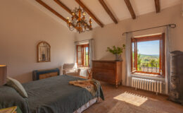 Cozy bedroom featuring a green bedspread, wooden furniture, and a chandelier, with natural light streaming through large windows that offer a view of the outdoors. The room has exposed wooden beams and a warm, inviting atmosphere.