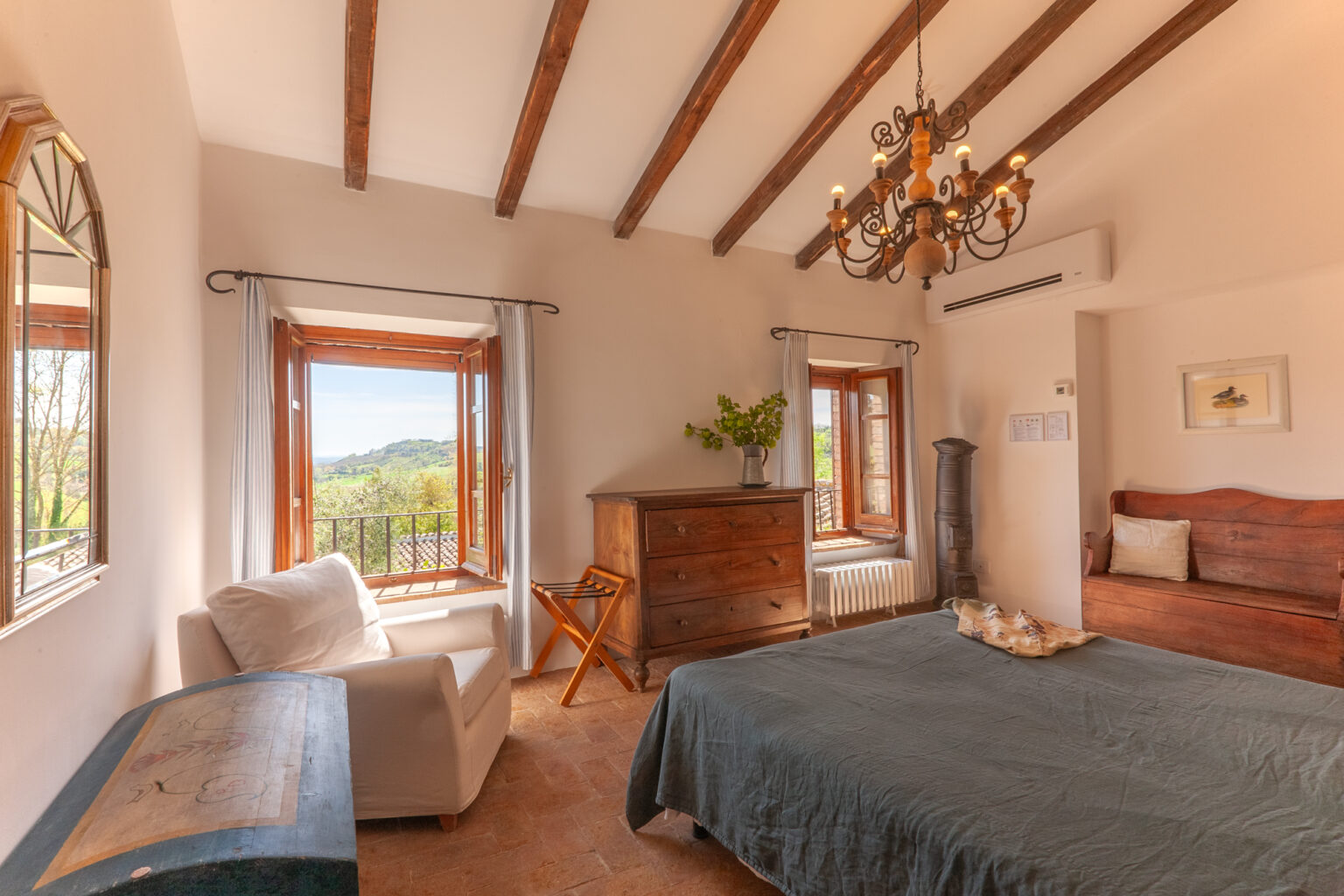 Cozy bedroom with wooden beams, featuring a comfortable bed, vintage dresser, and large windows offering scenic views. The room is decorated with a chandelier and a potted plant, creating a warm and inviting atmosphere. | Only on SopranoVillas